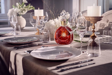 Bottle of Louis XIII cognac placed among crystal glasses, a candle on a wooden stand and a bouquet of flowers on a festive arrangement